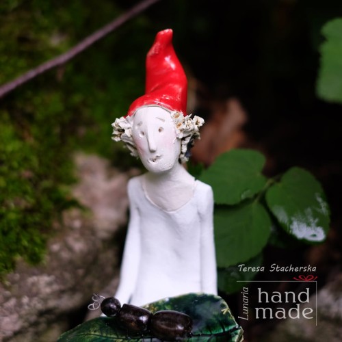 Ceramiczna figurka wesołego  leśnego skrzata w pozycji siedzącej trzymającego na kolanach zielony liść z czarną mrówką.jpg